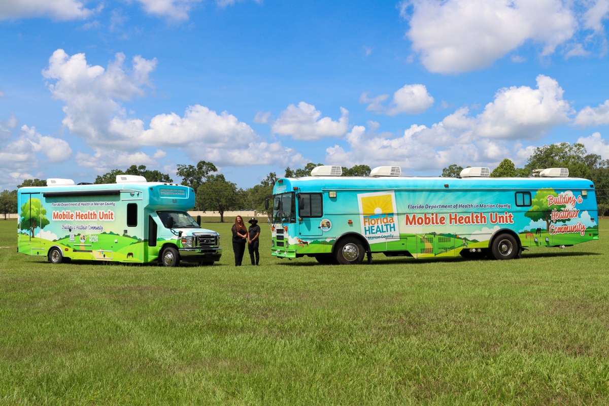 The Mini-MHU, two MHU staff members, and the main Mobile Health Unit in a grassy field.